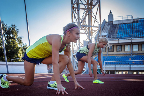 Δύο γυναικεία sprinter αθλητής ετοιμάζεται να ξεκινήσει μια κούρσα στο κόκκινο στίβου σε στάδιο στίβου. - Φωτογραφία, εικόνα