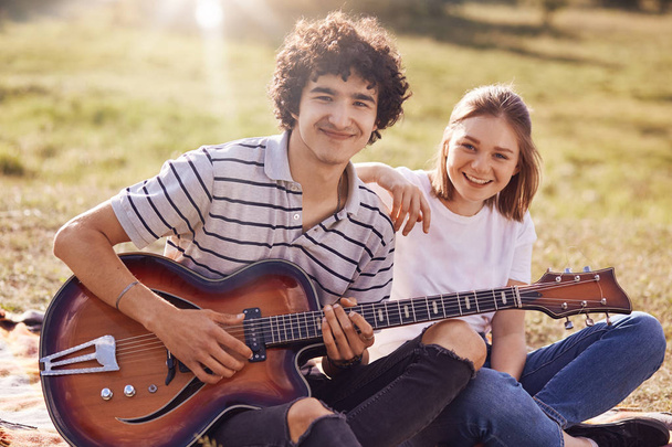 Two happy friends have joyful expression, gentle smiles on faces, recreat during summer time outdoor, play guitar and sing popular songs, can`t imagine their lives without music. Rest concept - Fotó, kép