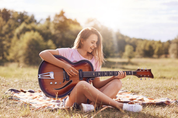 Beautiful young smiling female plays acoustic guitar, sits cross legs on green grass, being in good mood, learns new song, has appealing appearance. People, talent, youth and recreation concept - Foto, Imagem