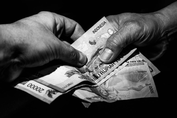 Twee handen, een Indonesisch en één van de Kaukasische wit overhandigen Indonesische bankbiljetten aan elkaar. Concept van het kopen van drugs door toeristen, buitenlandse investeringen in Indonesië, omkopen.  - Foto, afbeelding