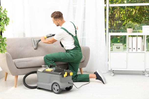 Male janitor removing dirt from sofa with upholstery cleaner in room - 写真・画像
