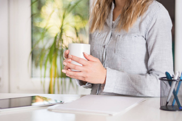 Business woman sitting on desk at modern office and holding white cup of coffee in hand. Girl wears elegant shirt on strips. Blurred big green windows in background. Close up, selective focus - Photo, Image