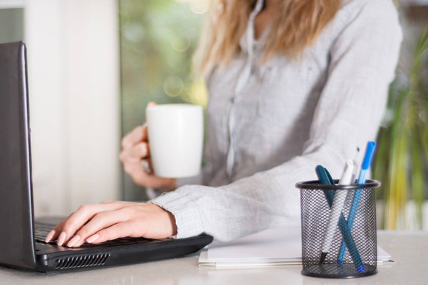 Деловая женщина в современном офисе работает над ноутбуком и держит чашку кофе в руке. Девушка носит элегантную рубашку и сидит на столе, размытые большие окна на заднем плане. Крупный план, избирательный фокус
 - Фото, изображение