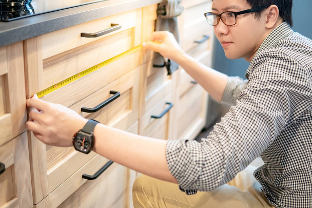 Joven hombre asiático usando cinta métrica para medir cajón en el mostrador de cocina de madera en el showroom. Compras de muebles para la mejora del hogar. Concepto de diseño interior
 - Foto, imagen