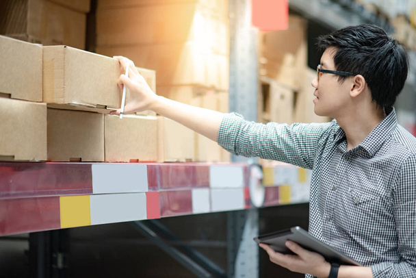 Jonge Aziatische man werknemer doet inventarisatie van het product in kartonnen doos op planken in magazijn met behulp van digitale tablet en pen. Concept van de feitelijke inventarisatie - Foto, afbeelding