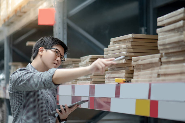 Молодой азиатский работник делает склад продукции в картонной коробке на полках на складе с помощью цифровых планшетов и ручки. Концепция подсчета физических запасов
 - Фото, изображение