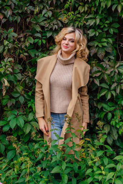 Young beautiful woman in sweater and coat stands next to the background of wild grapes leaves in autumn park - Foto, Imagen