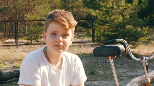 Een tiener jongen zit in bos zomer park naast een fiets en kijkt naar de camera - Video