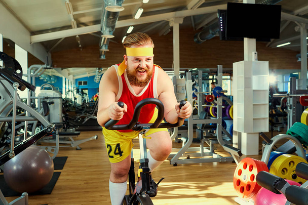 A funny fat man  doing exercises on an exercise bike in a sports - Valokuva, kuva