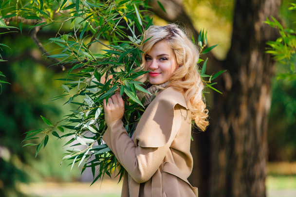 Beautiful elegant blonde woman dressed in a coat standing under the tree in autumn park. Sunny october day. - Foto, imagen