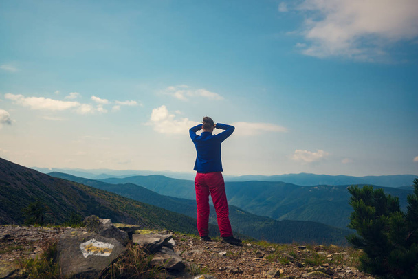 Gelukkige vrouw, wandelaar staat op een rotsachtige bergkam op een zonnige dag, bewondert de adembenemend uitzicht op de groene bergketens die zich uitstrekt tot de blauwe horizon. Epische reis door de wildernis. Achteraanzicht.  - Foto, afbeelding