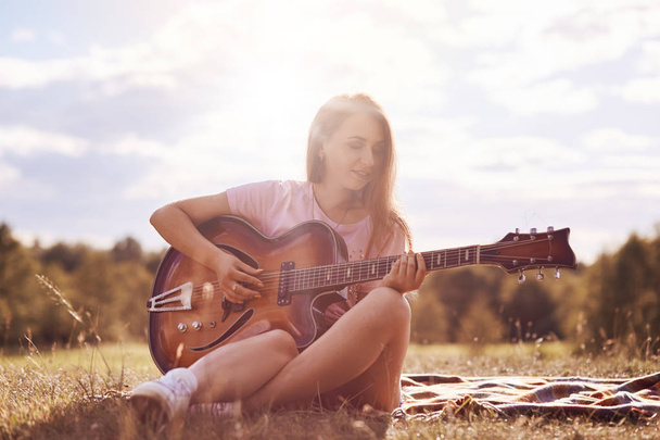 Attractive young female guitarist uses music instrument, plays guitar, sings song, produces wonderful sound or melody, sits crossed legs on plaid, poses against blue sky and sunshine background - Foto, Bild