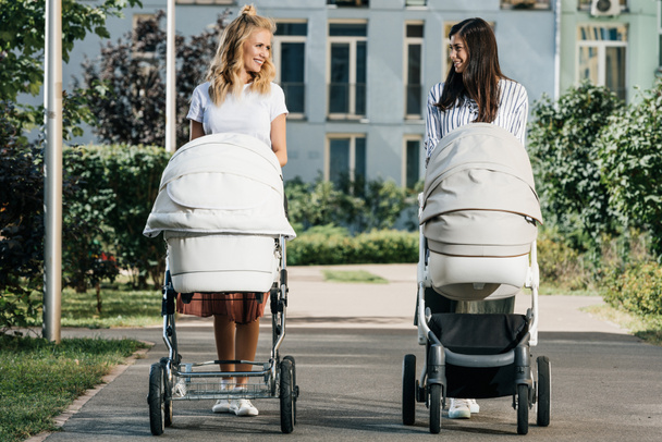 Mütter, die mit Kinderwagen auf der Straße spazieren gehen und einander anschauen  - Foto, Bild