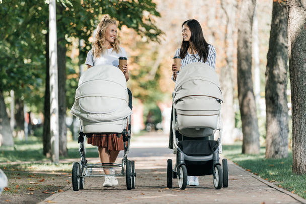 Mütter, die mit Kinderwagen und Coffee to go im Park spazieren gehen und einander anschauen  - Foto, Bild