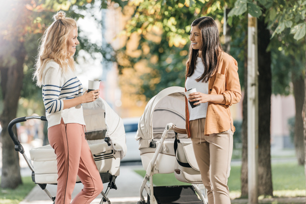 Μητέρες μιλούν, στέκεται με καφέ να πάει και το μωρό καροτσάκια στο πάρκο  - Φωτογραφία, εικόνα