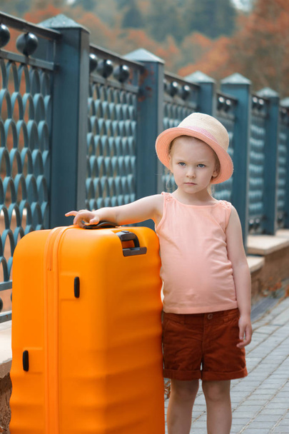 Pieni tyttö seisoo ison oranssin matkalaukun vieressä. Kaunis muotokuva lapsesta. Matkailun aihe
. - Valokuva, kuva