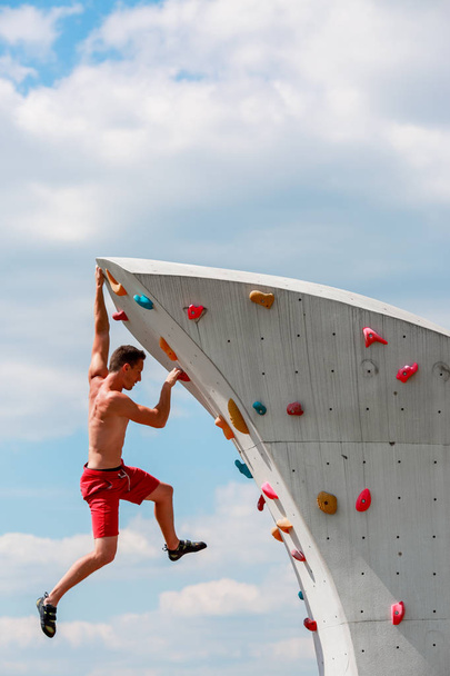 Photo of trained sports brunet in red shorts hanging on wall for rock climbing against blue sky with clouds - Foto, afbeelding