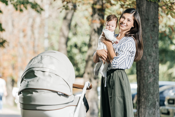 smiling mother holding baby near stroller in park and looking at camera - Photo, Image