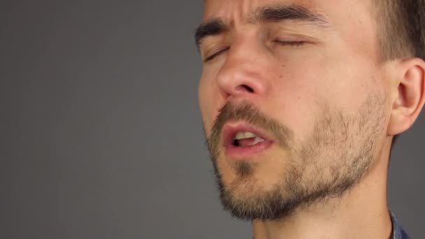 Portret van een bebaarde man niest en veegt zijn lippen met zijn hand, zijaanzicht, grijze achtergrond - Video