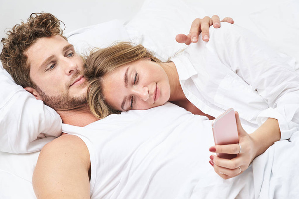 Young woman using Smartphone in bed with boyfriend - 写真・画像