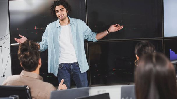 glimlachend jonge zakenman met brede armen presentatie tonen aan zakelijke partners op moderne kantoor  - Foto, afbeelding