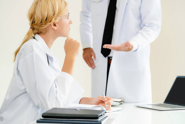Young female doctor at hospital office having conversation talking with another male doctor standing beside the table. Concept of medical healthcare professional team. - Photo, Image