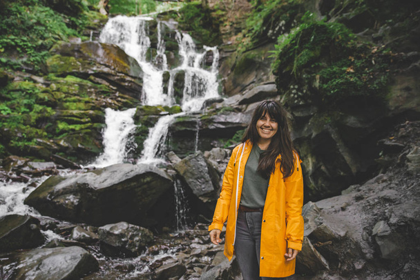 woman standing in yellow raincoat and looking at waterfall. hiking concept - Photo, Image