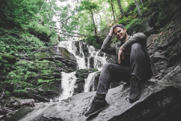 Mies istuu kivellä vesiputous taustalla. matkustuskäsite
 - Valokuva, kuva