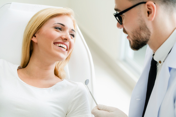 Молодой красивый стоматолог беседует со счастливой пациенткой, сидящей на стоматологическом стуле в стоматологической клинике. Концепция стоматологии
. - Фото, изображение