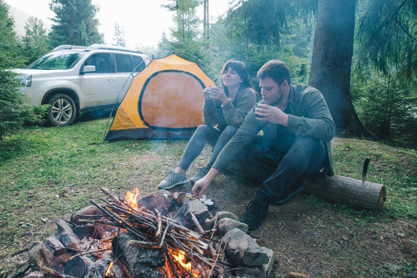 couple sitting near camp fire and drinking tea. tent and suv on background. union with nature - Fotografie, Obrázek
