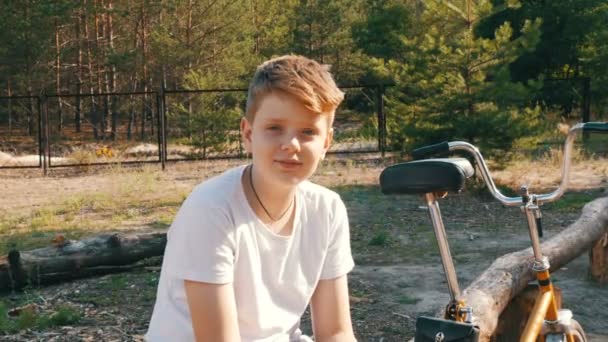Aranyos szőke, kék szemek egy tinédzser fiú ül, és mosolyog egy parkban, kerékpár és a tárgyalások mellett nyáron. Egy fiú érzelmi portréja - Felvétel, videó