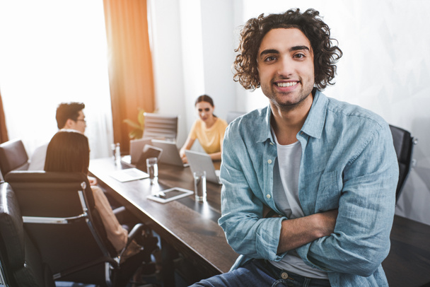 jeune homme d'affaires souriant avec les mains croisées regardant la caméra et ses partenaires travaillant derrière au bureau moderne
  - Photo, image