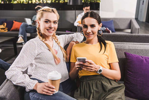 two smiling businesswomen with coffee and smartphone at modern coworking office  - Fotografie, Obrázek