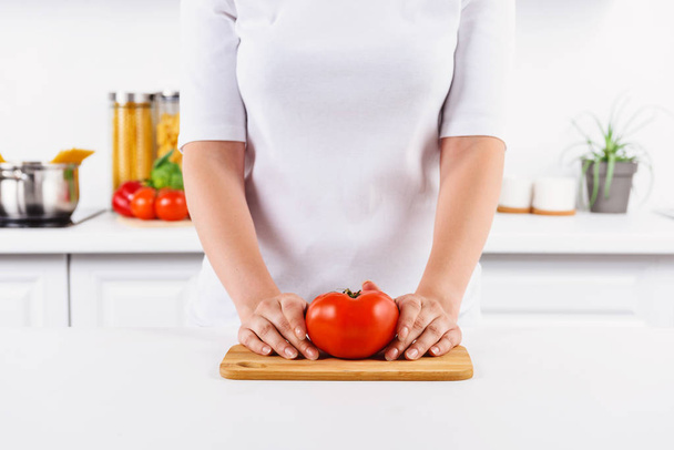 cropped image of woman holding ripe tomato on cutting board in light kitchen - Zdjęcie, obraz