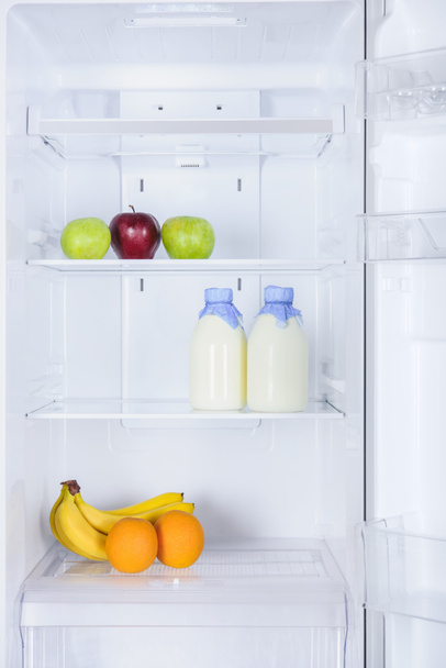 ripe tasty apples, bananas, oranges and bottles of milk in fridge - Photo, image