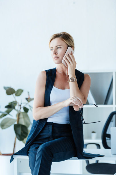 confident businesswoman holding eyeglasses and talking on smartphone in office - Photo, image
