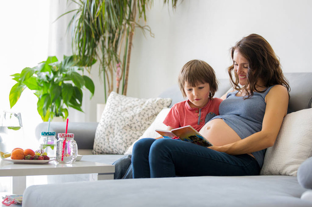 Young pregnant woman, reading a book at home to her boy, eating fruits, hugging and laughing - Φωτογραφία, εικόνα