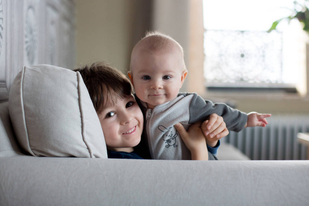 Little baby boy and his older brother, sitting on a couch in sunny living room, smiling with happiness - Foto, Imagem