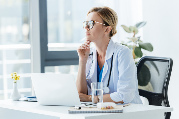 thoughtful female doctor in eyeglasses sitting at table with laptop in office  - Фото, изображение
