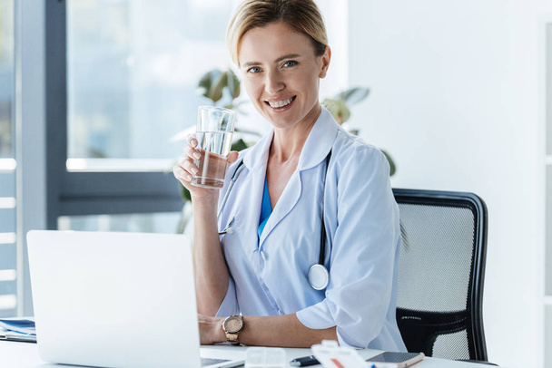 ευτυχισμένη γυναίκα γιατρό κρατώντας το ποτήρι νερό και να κάθεται στο τραπέζι με το laptop στο γραφείο  - Φωτογραφία, εικόνα