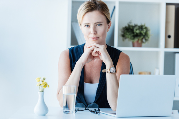 portrait of adult businesswoman looking at camera and sitting at table with laptop in office  - Photo, image