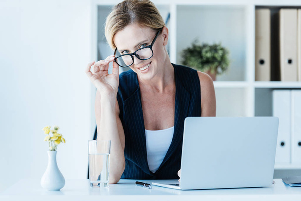 portrait of smiling businesswoman in eyeglasses looking at camera while sitting at table with laptop in office  - Foto, Imagem