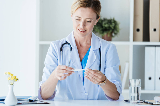adult female doctor in white coat checking thermometer at table in office  - Photo, Image