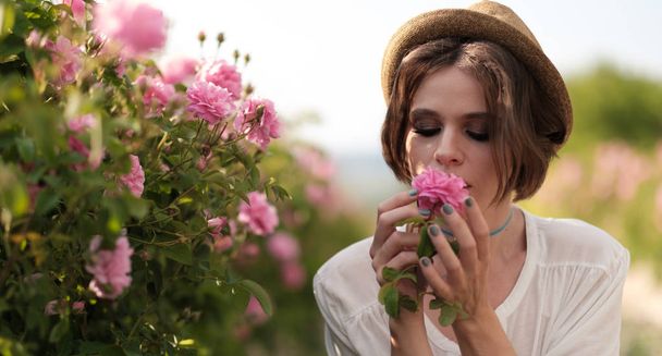 Bella giovane donna con i capelli ricci posa vicino a rose in un giardino. Il concetto di pubblicità dei profumi
. - Foto, immagini