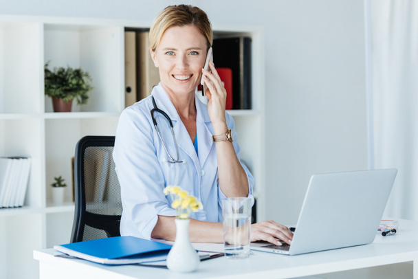 happy female doctor looking at camera and talking on smartphone at table with laptop in office  - Photo, Image