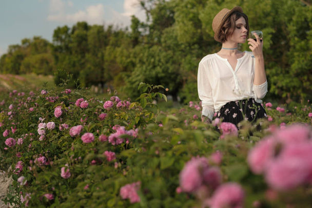 Όμορφη νεαρή γυναίκα με σγουρά μαλλιά ποζάρει κοντά τριαντάφυλλα μέσα σε έναν κήπο. Κορίτσι με ένα ποτήρι στο χέρι, δοκιμάζοντας κόκκινο κρασί και διασκέδαση - Φωτογραφία, εικόνα