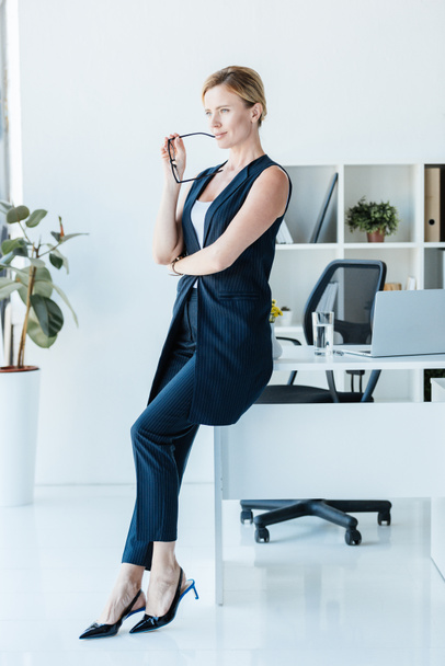 thoughtful businesswoman holding eyeglasses near table with laptop in office  - Фото, зображення