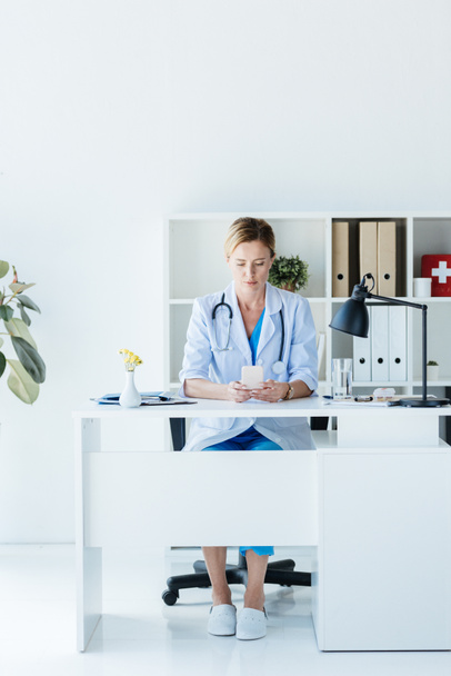 volwassen vrouwelijke arts in de witte jas met smartphone aan tafel in kantoor  - Foto, afbeelding