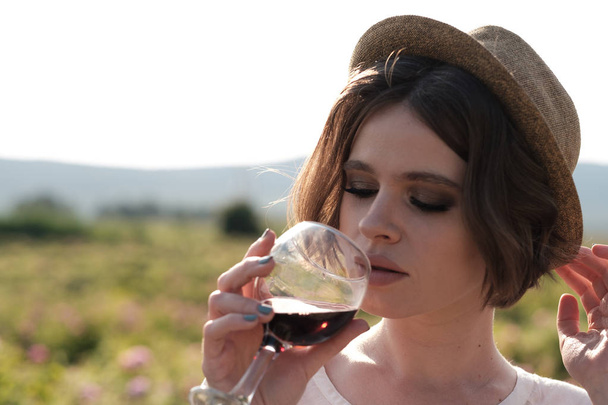 庭のバラに近いポーズ巻き毛の美しい若い女性。赤ワインを試飲し、楽しい時を過す彼女の手でガラスを持つ少女 - 写真・画像