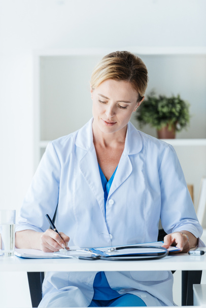 fokussierte Ärztin in weißem Mantel mit Klemmbrett am Tisch im Büro  - Foto, Bild